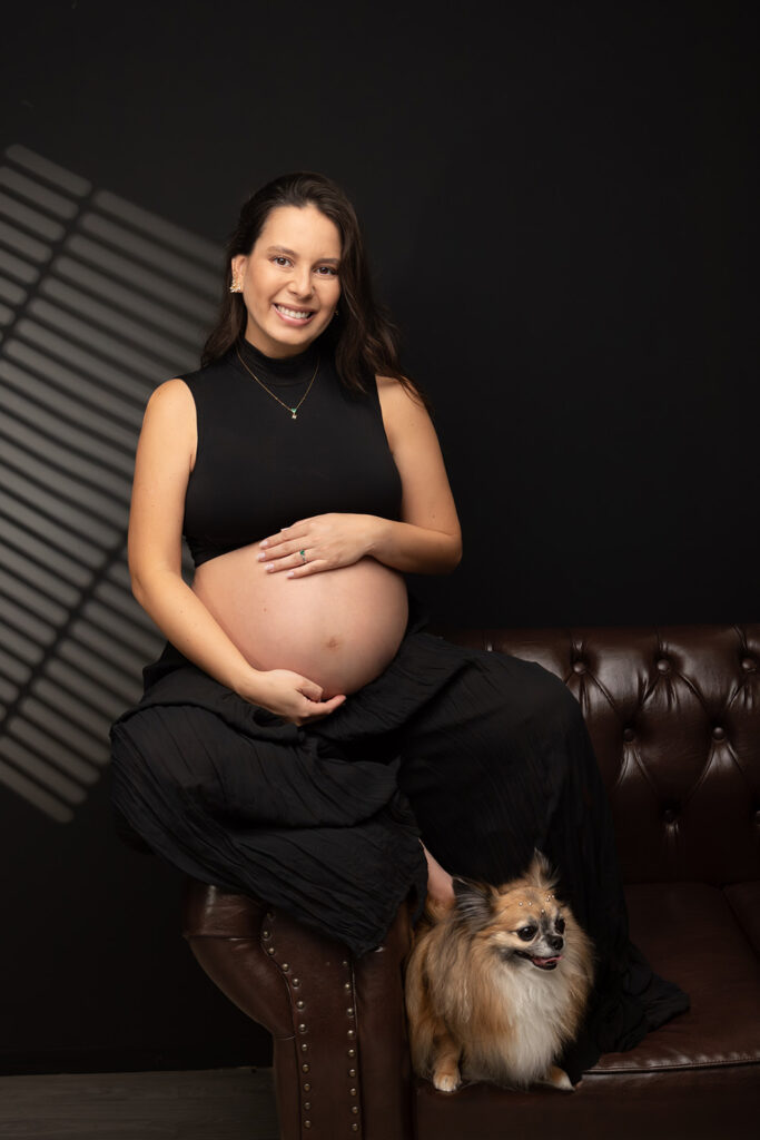 Imagem de gestante sorridente com barriga à mostra, sentada em sofá com seu cachorro, em estúdio com.