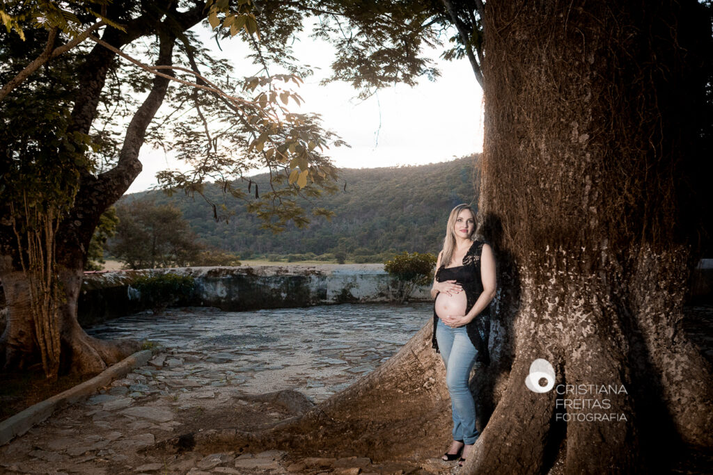 Mulher grávida posando ao lado de árvore em cenário natural externo.