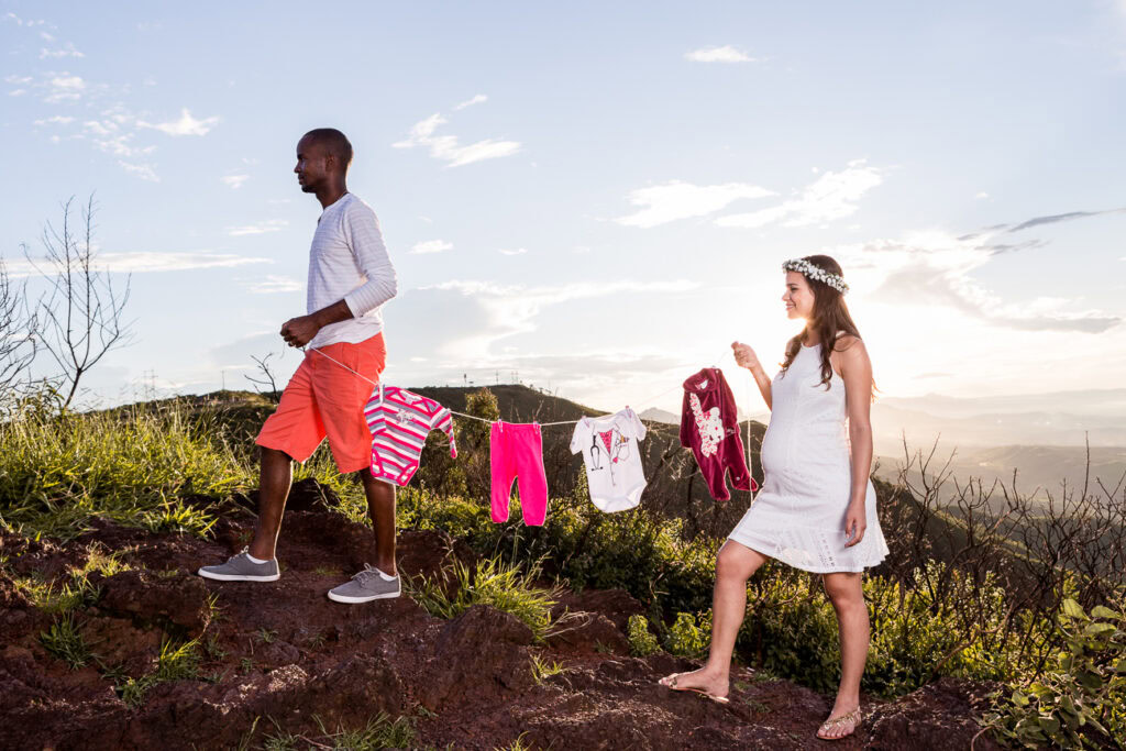 Imagem de gestante e acompanhante ao ar livre, com roupas leves, em cenário natural ao pôr do sol.
