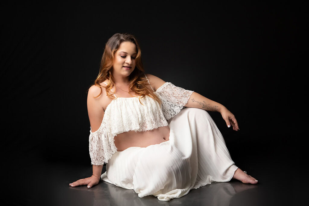 Foto de gestante em ensaio de maternidade com vestido branco e fundo escuro.