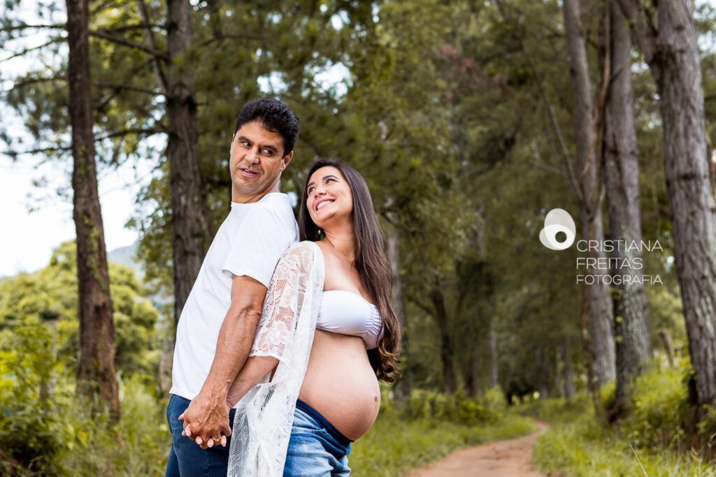 Retrato de casal grávido ao ar livre, com árvores ao fundo, durante sessão de fotos de gestante.